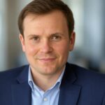 Markus Becker Porträtfoto von Markus Becker. Er ist ein junger Mann mit kurzen, braunen Haaren und einem freundlichen Lächeln. Er trägt einen dunkelblauen Anzug mit einem hellblauen Hemd. Der Hintergrund ist modern und leicht unscharf, was eine Büroumgebung suggeriert.